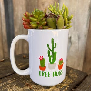 May include: White ceramic mug with a green cactus design and the text "FREE HUGS". The mug is filled with a small succulent plant.
