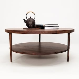 May include: A round wooden coffee table with a lower shelf. The table has a dark brown finish and is made of solid wood. A teapot and teacups are on the top shelf, and a stack of books is on the table.