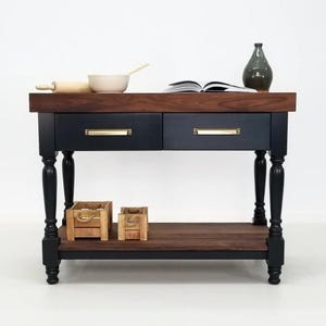 May include: A kitchen island with a dark walnut wood top and a black base featuring two drawers with brass-toned handles. The island has four turned legs, a lower shelf, and two small wooden crates on the bottom shelf. The top surface holds a bowl.