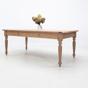 May include: A light brown wooden dining table with two drawers and turned legs. A clear glass vase with dried flowers sits on the table. The table is a light natural wood color and is set against a white background.