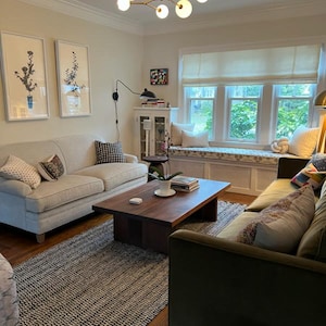 K&ouml;nnte beinhalten: Ein Wohnzimmer mit einem wei&szlig;en Sofa, einem gr&uuml;nen Sofa, einem Couchtisch, einem Teppich und einem Fenster mit einer wei&szlig;en Fensterbank. Der Raum ist mit Kunstwerken, Pflanzen und Kissen dekoriert.