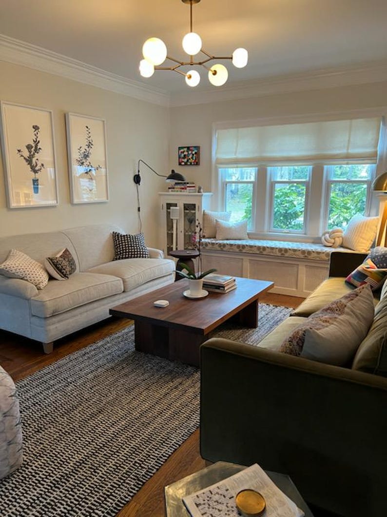 K&ouml;nnte beinhalten: Ein Wohnzimmer mit einem wei&szlig;en Sofa, einem gr&uuml;nen Sofa, einem Couchtisch, einem Teppich und einem Fenster mit einer wei&szlig;en Fensterbank. Der Raum ist mit Kunstwerken, Pflanzen und Kissen dekoriert.