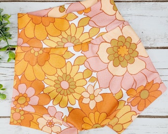 May include: A pair of pink and orange floral print baby bloomers. The bloomers have a wide elastic waistband and a ruffled hem.
