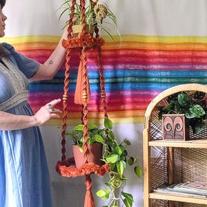 May include: A hanging macrame plant holder in shades of orange and brown, with two shelves holding potted plants. The plant holder is suspended from the ceiling. A woven wicker shelf unit is to the right, and a colorful rainbow backdrop is behind the plant holder.
