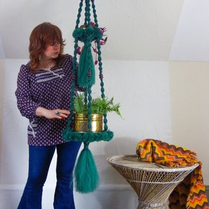 May include: A green macrame plant hanger with a gold pot and a green plant hanging from the ceiling. A woman is standing in front of the hanger wearing a blue and white patterned shirt and blue jeans. A wicker table with a brown and orange blanket is in the foreground.