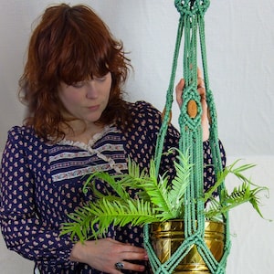 May include: A green macrame plant hanger with a wooden bead accent. The hanger is holding a brass pot with a green plant.