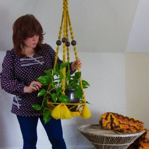 May include: A yellow macrame plant hanger with wooden beads hangs from the ceiling. A potted plant is in the hanger. A woman is holding the hanger and wearing a blue and black patterned blouse and blue jeans.