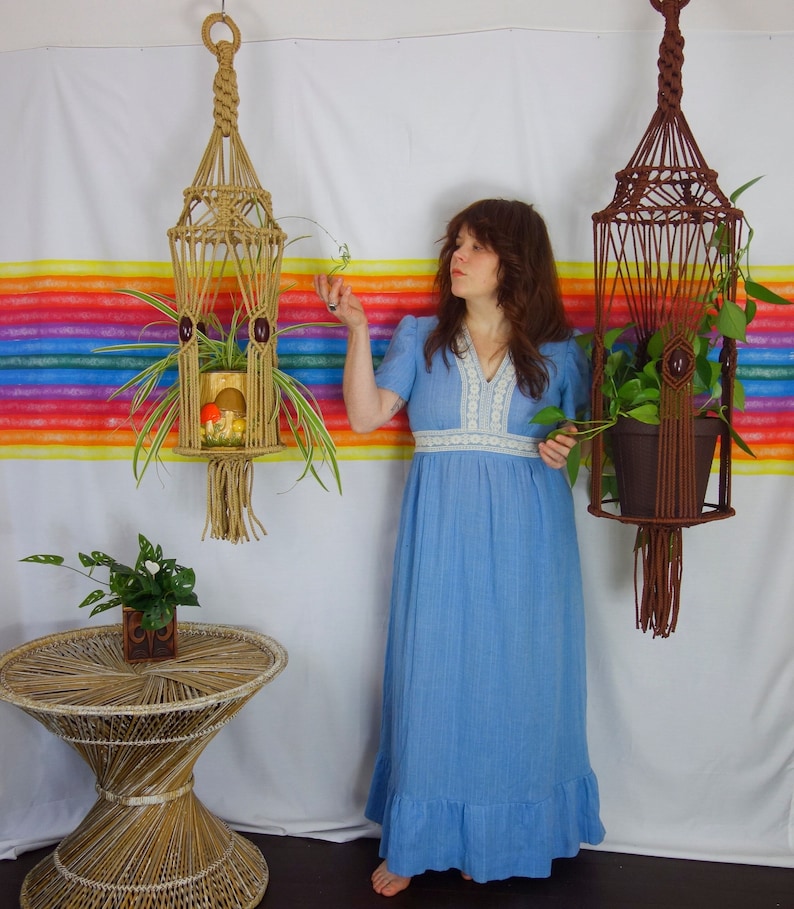 A woman in a long, vintage, sky blue dress stands between two  vintage-style handmade XL  plant hangers and tends to the plants they hold
