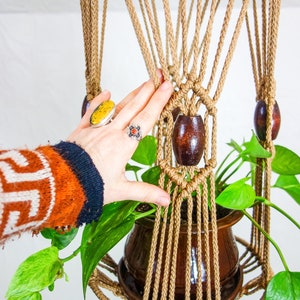 May include: A macrame plant hanger with a brown pot and green plant. The hanger is made of natural rope and has wooden beads.