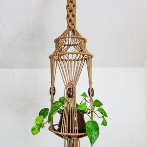 May include: A macrame plant hanger with a brown pot and green plant. The hanger has wooden beads and a fringe at the bottom.