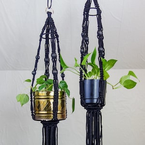 May include: Two black macrame plant hangers with gold and black pots. The hangers have black beads and tassels.