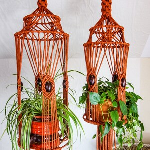 May include: Two macrame plant hangers with wooden beads, each holding a potted plant. The hangers are made of brown cord and have a geometric design.
