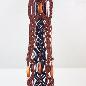 May include: A brown and black macrame wall hanging with wooden beads. The hanging is made with a knotting technique and has a geometric pattern.