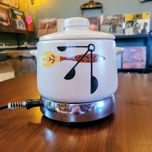 May include: A vintage, off-white electric fondue pot with a lid and side handles. The pot features a whimsical design of a yellow fork, a red fish, and black utensils. It sits on a chrome base with a power cord, perfect for entertaining.