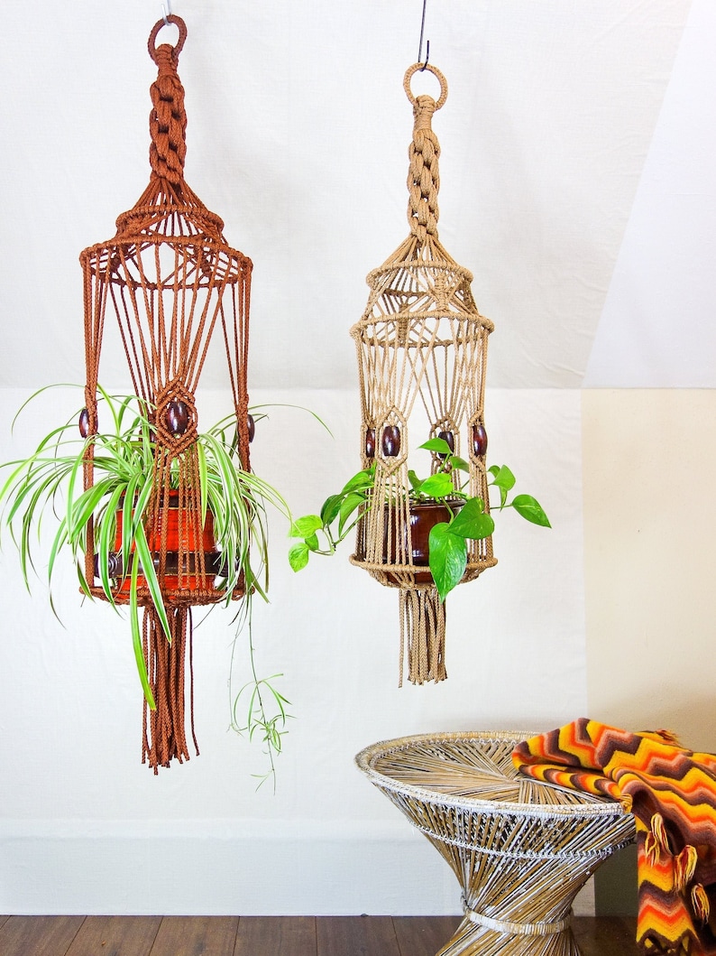 May include: Two macrame plant hangers with wooden beads, one brown and one beige, hanging from the ceiling with plants inside.