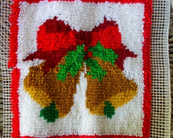 May include: A square, handmade Christmas decoration featuring a design of two golden bells and a red bow on a white background. The bells have green accents, and the border is red. The piece is made with a textured, looped yarn technique.