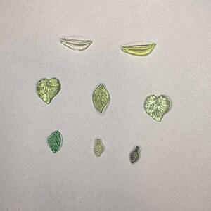 May include: A collection of seven small, translucent glass beads shaped like leaves and hearts in various shades of light green and yellow. The beads feature detailed textures and small holes for stringing, arranged in a scattered pattern.
