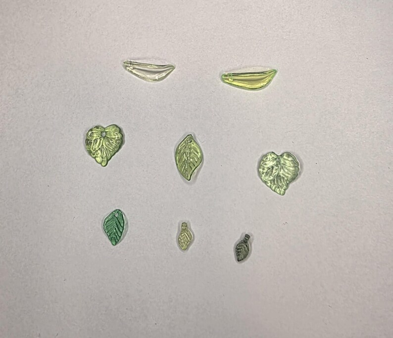May include: A collection of seven small, translucent glass beads shaped like leaves and hearts in various shades of light green and yellow. The beads feature detailed textures and small holes for stringing, arranged in a scattered pattern.