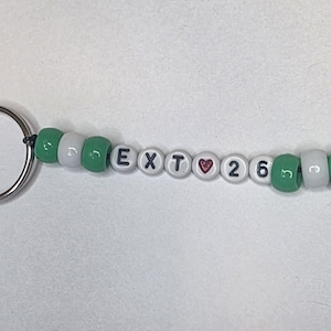 May include: A keychain featuring a silver metal lobster clasp and ring attached to a string of beads. The beads alternate between solid green and white, with white letter beads spelling EXT, a red heart bead, and the number beads 26, ending with a green leaf charm.