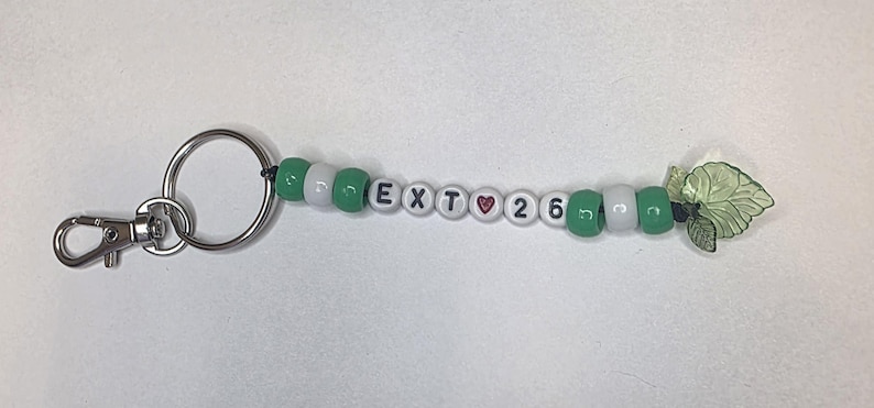 May include: A keychain featuring a silver metal lobster clasp and ring attached to a string of beads. The beads alternate between solid green and white, with white letter beads spelling EXT, a red heart bead, and the number beads 26, ending with a green leaf charm.