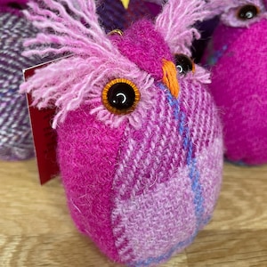 May include: A handmade, plush owl in shades of pink and purple tweed fabric. The owl has large, black button eyes with orange accents, and fluffy pink feather-like details. The owl is part of a collection of similar handcrafted items.