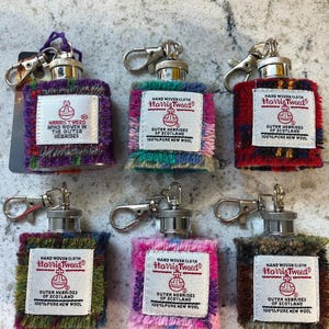 Op de afbeelding: Een verzameling Harris Tweed sleutelhangers in verschillende kleuren, waaronder paars, groen, rood en roze. Elke sleutelhanger heeft een metalen bovenkant en sluiting, met een geweven stoffen behuizing met het Harris Tweed label. De labels luiden "Hand Woven Cloth" en "Outer Hebrides of Scotland."