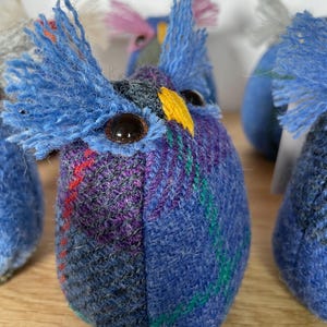 May include: Handmade owl-shaped plush toys. The owls are made of blue and purple plaid fabric with tufted blue feather accents. Each owl has large, dark brown eyes and a yellow beak. The owls are displayed on a wooden surface.