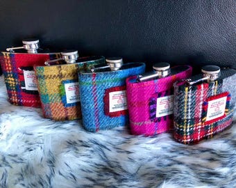 May include: A collection of six hip flasks, each wrapped in colorful tweed fabric. The flasks feature a variety of plaid patterns in red, blue, yellow, pink, and gray. Each flask has a silver cap and a small label.