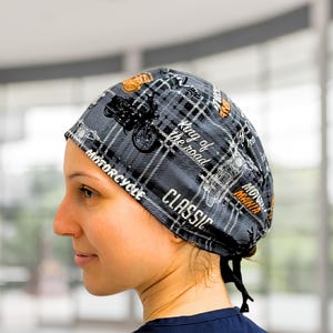 May include: A grey and black plaid scrub cap featuring a motorcycle-themed print with text including "King of the road," "MOTORCYCLE," "CLASSIC," and "MOTOR MANIA." The cap has a tie-back closure and is worn over the head with a dark blue shirt.