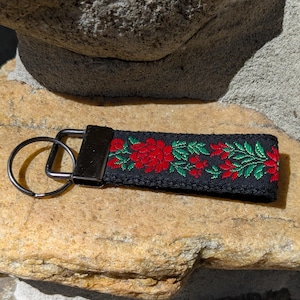 May include: Black fabric keychain with a floral design. The keychain features a rectangular black fabric strap with a metal clasp and key ring. The strap is embroidered with red flowers and green leaves.
