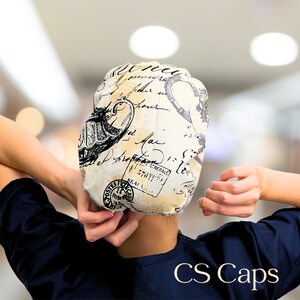 May include: A cream-colored cap with a vintage script and stamp design. The cap has black and white accents and is being held up by a person. The text "CS Caps" is at the bottom of the image.