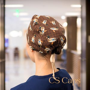 May include: Brown duck print scrub cap. The cap features a repeating pattern of ducks in shades of brown, blue, and white. The cap is tied in the back with a beige ribbon. The text "CS Caps" is visible on the bottom right.