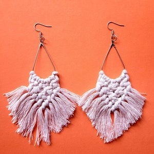 Peut inclure: Une paire de boucles d'oreilles macramé faites à la main. Elles présentent un motif tissé blanc cassé avec un cadre triangulaire en fil de fer et de longues franges. Les boucles d'oreilles sont suspendues à des crochets d'oreilles argentés sur fond orange.