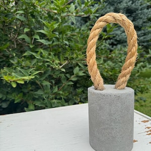 Peut inclure: Un poids cylindrique en béton gris doté d'une épaisse poignée en corde de jute beige en forme de boucle, solidement insérée dans la surface supérieure. L'objet présente une texture lisse et solide ainsi qu'une base robuste et une anse en fibre naturelle.