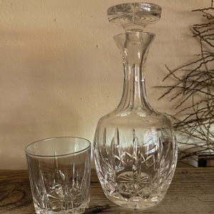 May include: A clear glass decanter with a matching stopper and a single glass tumbler. Both pieces feature a cut glass pattern with vertical lines and starburst motifs at the base, creating a reflective surface on the transparent glass.