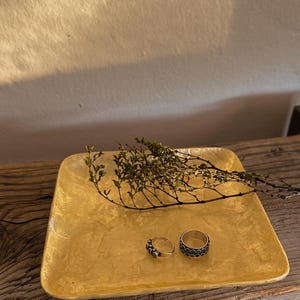 May include: A square, textured gold-colored tray holding two silver rings and a small, dried leafy branch. One ring features a thin band with small green accents, while the other is a wider band with a repeating circular pattern and dark inlays.
