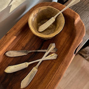 May include: A set of four silver-toned metal butter knives with decorative shell-like detailing on the handles, accompanied by a small, round, speckled tan ceramic bowl. The items are arranged on a dark, polished wooden serving tray.