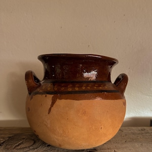 Op de afbeelding: Een ronde terracotta pot met twee zijhandvatten, voorzien van een brede opening en een glanzend donkerbruin geglazuurd bovengedeelte. Het onderste gedeelte van de pot is ongeglazuurd met een natuurlijke, matte, lichtbruine afwerking.