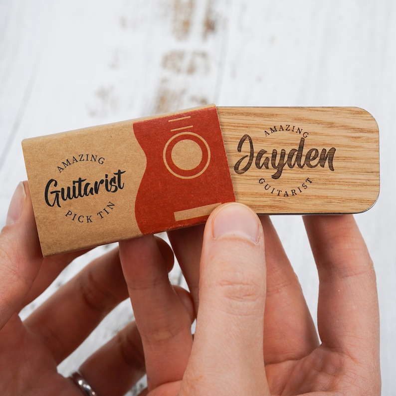 May include: A wooden guitar pick tin with the text "Amazing Guitarist Pick Tin" on the front. The tin is open and reveals a wooden guitar pick with the text "Amazing Jayden Guitarist" on the front.