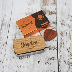May include: A wooden guitar pick tin with a personalized inscription that reads "Amazing Jayden Guitarist". The tin is closed and sitting on a white wooden surface. A wooden guitar pick is lying next to the tin.