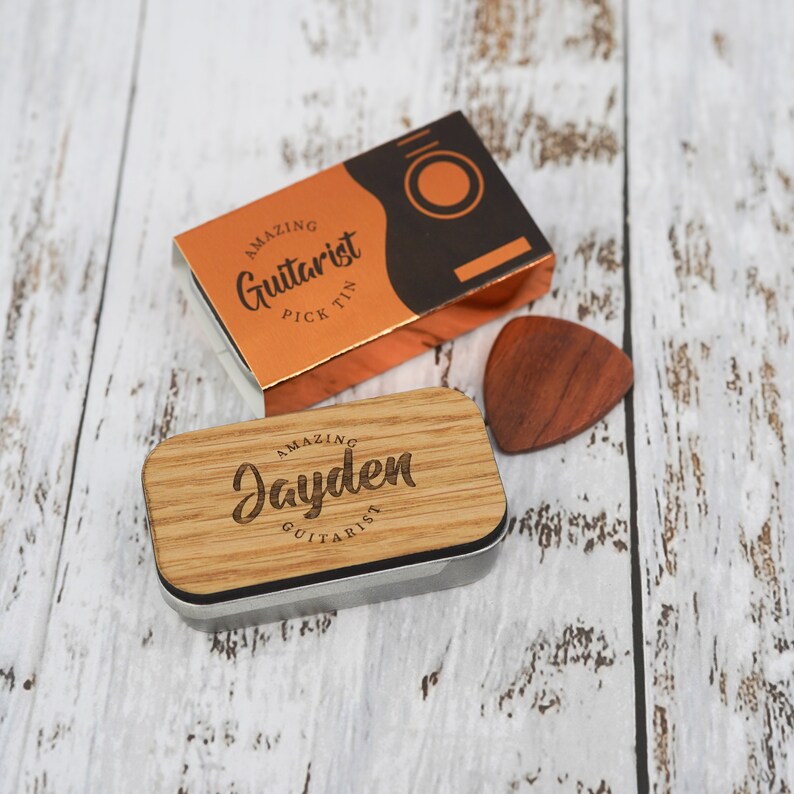 May include: A wooden guitar pick tin with a personalized inscription that reads "Amazing Jayden Guitarist". The tin is closed and sitting on a white wooden surface. A wooden guitar pick is lying next to the tin.