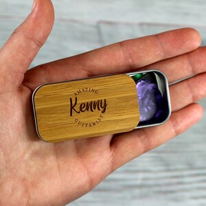 May include: A wooden guitar pick tin with the text "Amazing Kenny Guitarist" engraved on the lid. The tin is open and a purple guitar pick is visible inside.