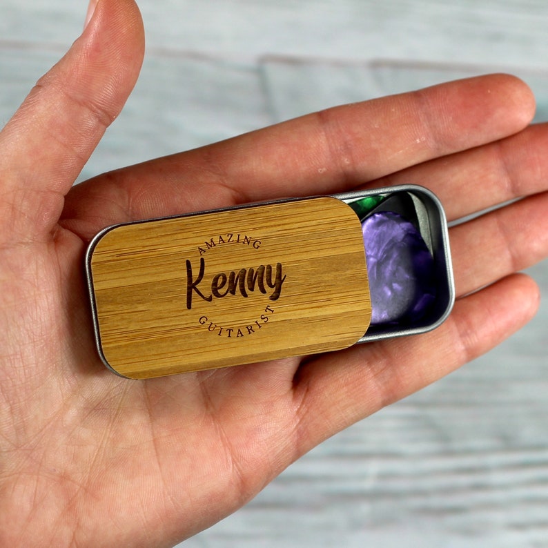 May include: A wooden guitar pick tin with the text "Amazing Kenny Guitarist" engraved on the lid. The tin is open and a purple guitar pick is visible inside.