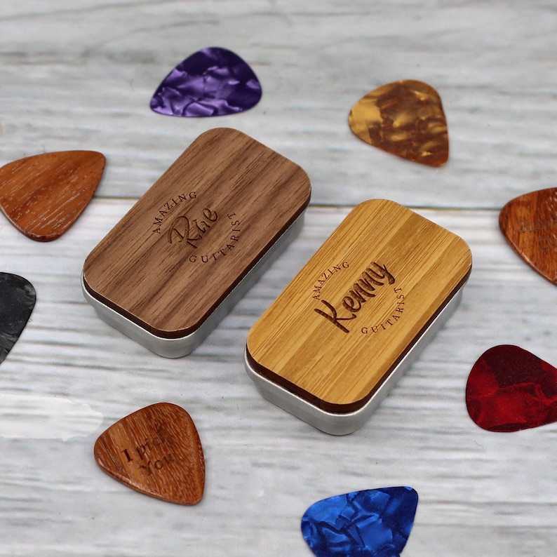 May include: Two wooden boxes with engraved text, "Amazing Rue Guitarist" and "Amazing Kenny Guitarist", are shown on a light wood surface. The boxes are open and contain guitar picks. There are also several guitar picks scattered around the boxes.