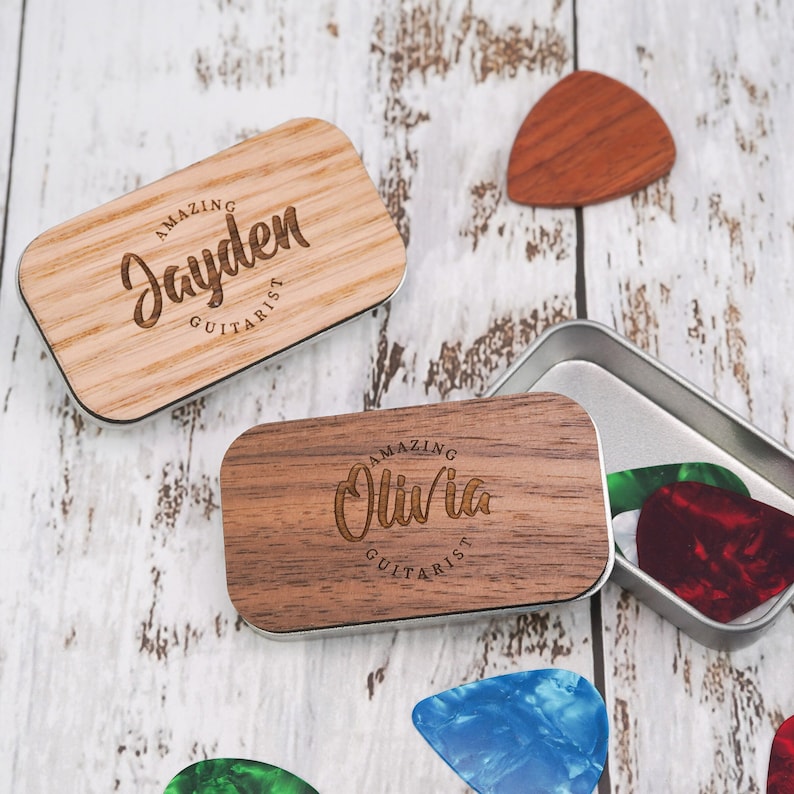 May include: Two wooden guitar pick holders with engraved names and the text "Amazing Guitarist". One holder is brown and the other is light brown. The holders are next to a metal tin with guitar picks inside.