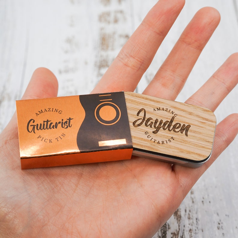 May include: A wooden guitar pick tin with a copper colored lid. The lid has the text "Amazing Guitarist Pick Tin" and a graphic of a guitar. The wooden part of the tin has the text "Amazing Jayden Guitarist".