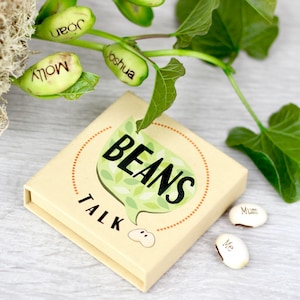 Peut inclure: Une petite boîte brune avec les mots "Beans Talk" imprimés dessus. La boîte a une feuille verte avec le mot "Beans" imprimé dessus. Il y a aussi plusieurs petites fèves blanches avec des noms écrits dessus, comme "Molly", "Joshua" et "Mum".