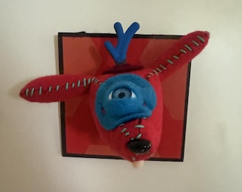 Op de afbeelding: Een rode vilten wezenkop gemonteerd op een vierkante rode plaat. Het wezen heeft één groot blauw oog, blauwe geweien en lichtgroene stiksels langs de oren en snuit. Het heeft een zwarte neus en een klein wit tongetje dat uitsteekt.