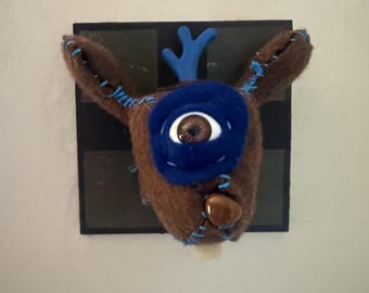 May include: A brown felted creature head with a single large eye, blue velvet accents, and blue stitched details. The creature has a blue antler-like protrusion on top and a brown bead nose, all mounted on a square black base for wall display.