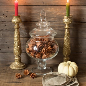 May include: An autumnal still life arrangement. A glass jar filled with pine cones sits between two gold candle holders with lit candles. A small white pumpkin and a linen napkin are also present.